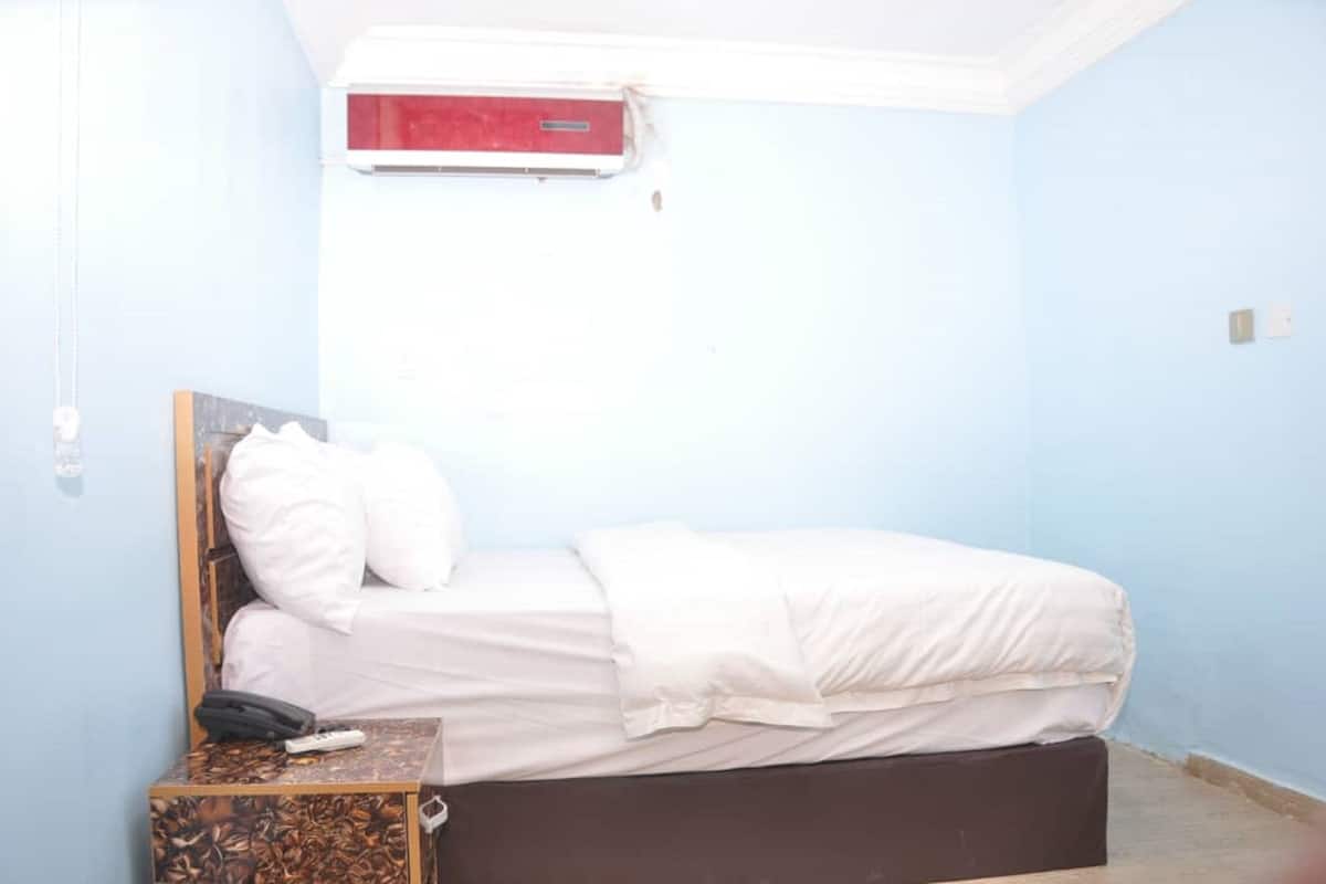 A single bed with a headboard, white bedding, and a small bedside table with a telephone and a remote control.
