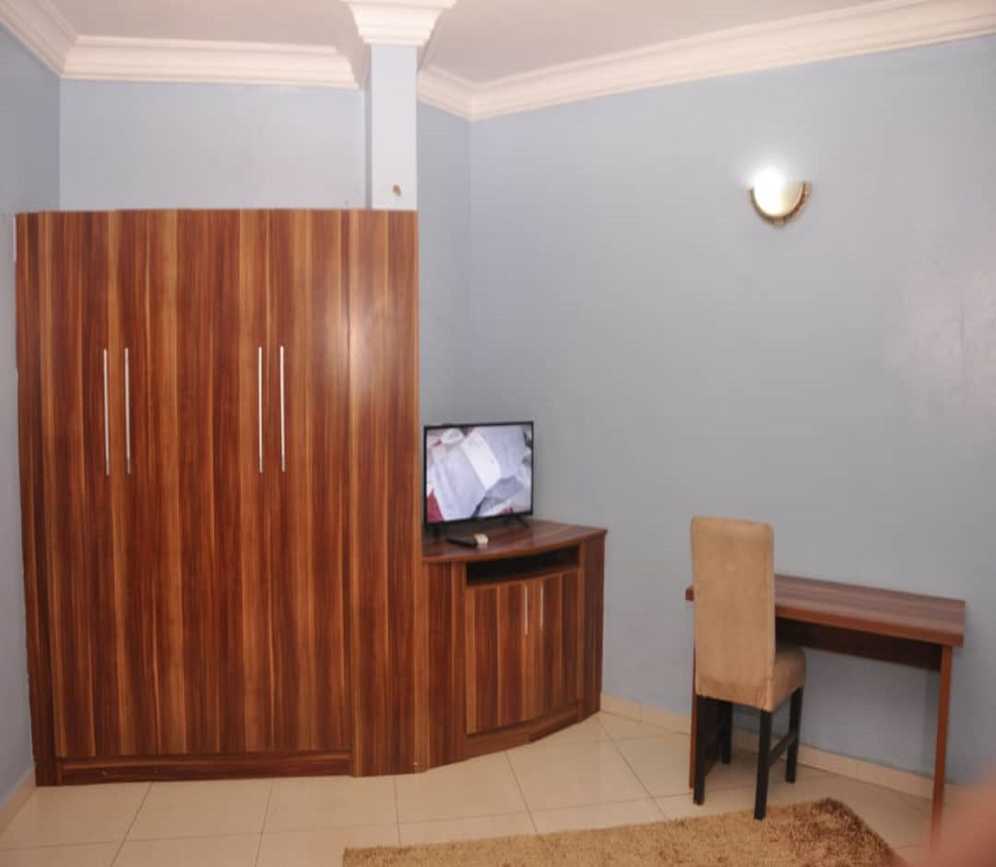 A room with a wooden wardrobe, a television on a wooden stand, a desk, and a chair.