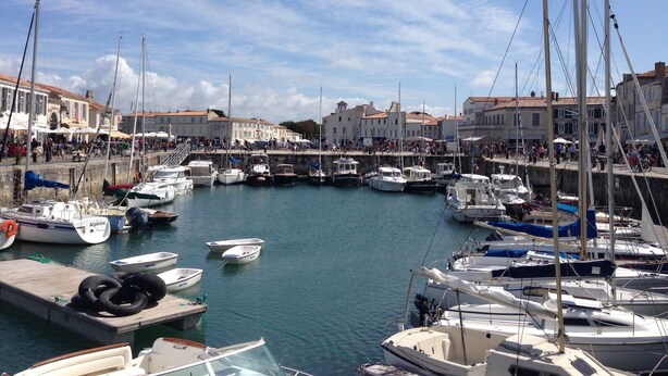 Un port de plaisance avec des bateaux amarrés, une promenade en bord de mer et des bâtiments aux toits recouverts de tuiles rouges.