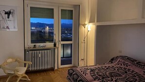 A bedroom with a bed, a radiator, a chair, and a window with a view of a cityscape at dusk. - Camilla 's House 2 - “it's cool“. (Torino)
