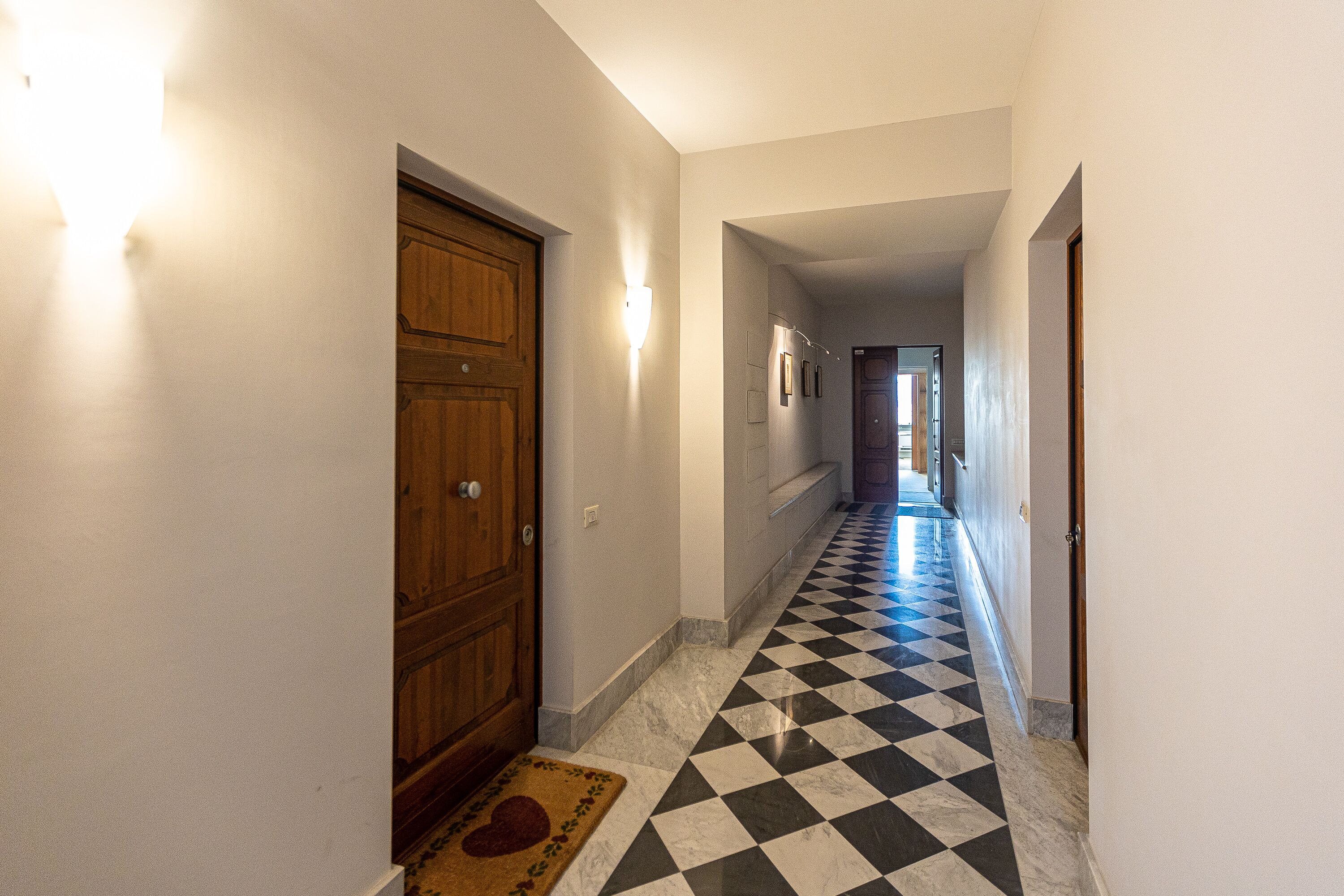 Un corridoio con pavimento a scacchi, porte in legno e lampade a parete.