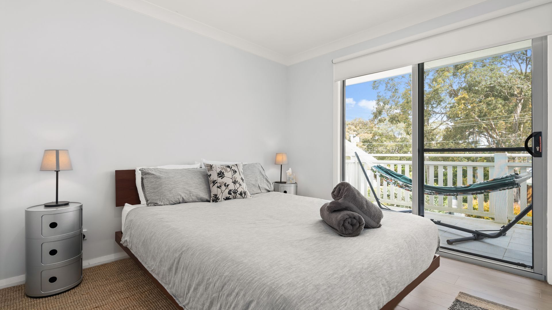 A bedroom with a large bed, two bedside lamps, a view of a balcony with a hammock, and a window with a view of trees.