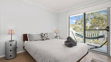A bedroom with a large bed, two bedside lamps, a view of a balcony with a hammock, and a window with a view of trees.