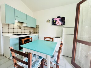 A compact kitchen with teal cabinets, a white countertop, and a dining table with chairs. A television is mounted on the wall, and a refrigerator is visible in the background.
