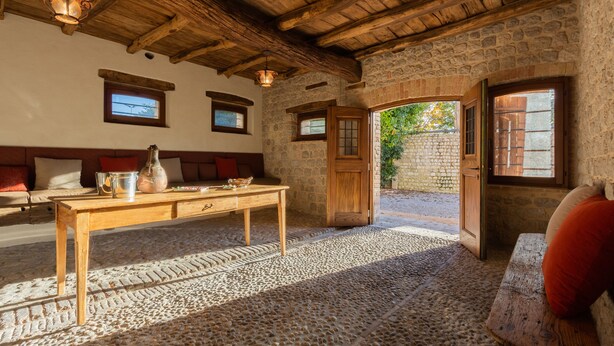 Una habitación rústica con pared de piedra, vigas de madera en el techo, una mesa de madera con un florero y un banco con cojines.