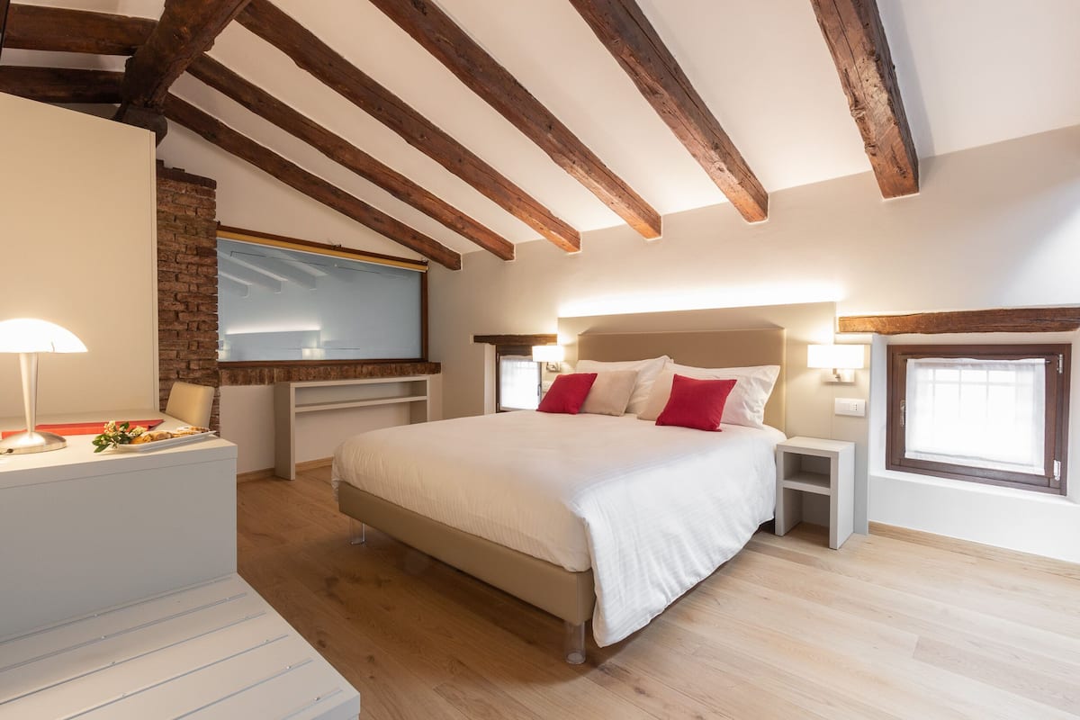 Une chambre avec un grand lit, des poutres en bois, un mur en briques et une fenêtre.