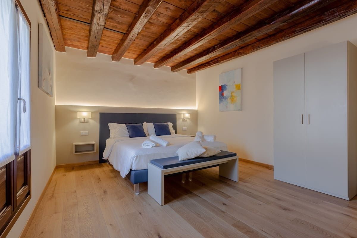 Une chambre avec un plafond en bois, un lit recouvert de draps blancs, un banc, une armoire et un luminaire fixé au mur.
