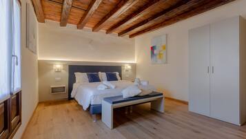 A bedroom with a wooden ceiling, a bed with white linens, a bench, a wardrobe, and a wall-mounted light fixture.