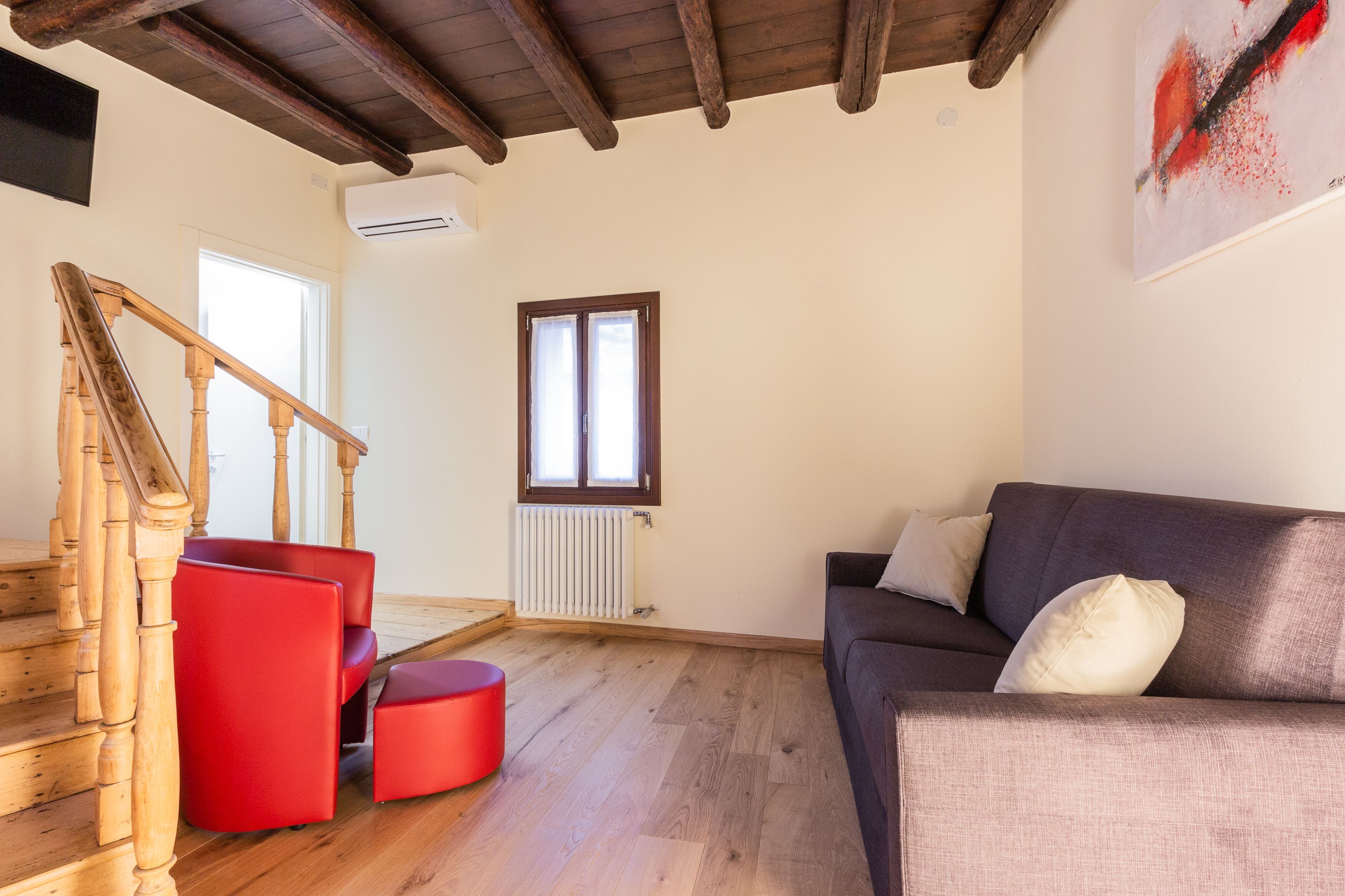 Una sala de estar con una escalera de madera, un sofá rojo, un sofá gris y una ventana con persianas.