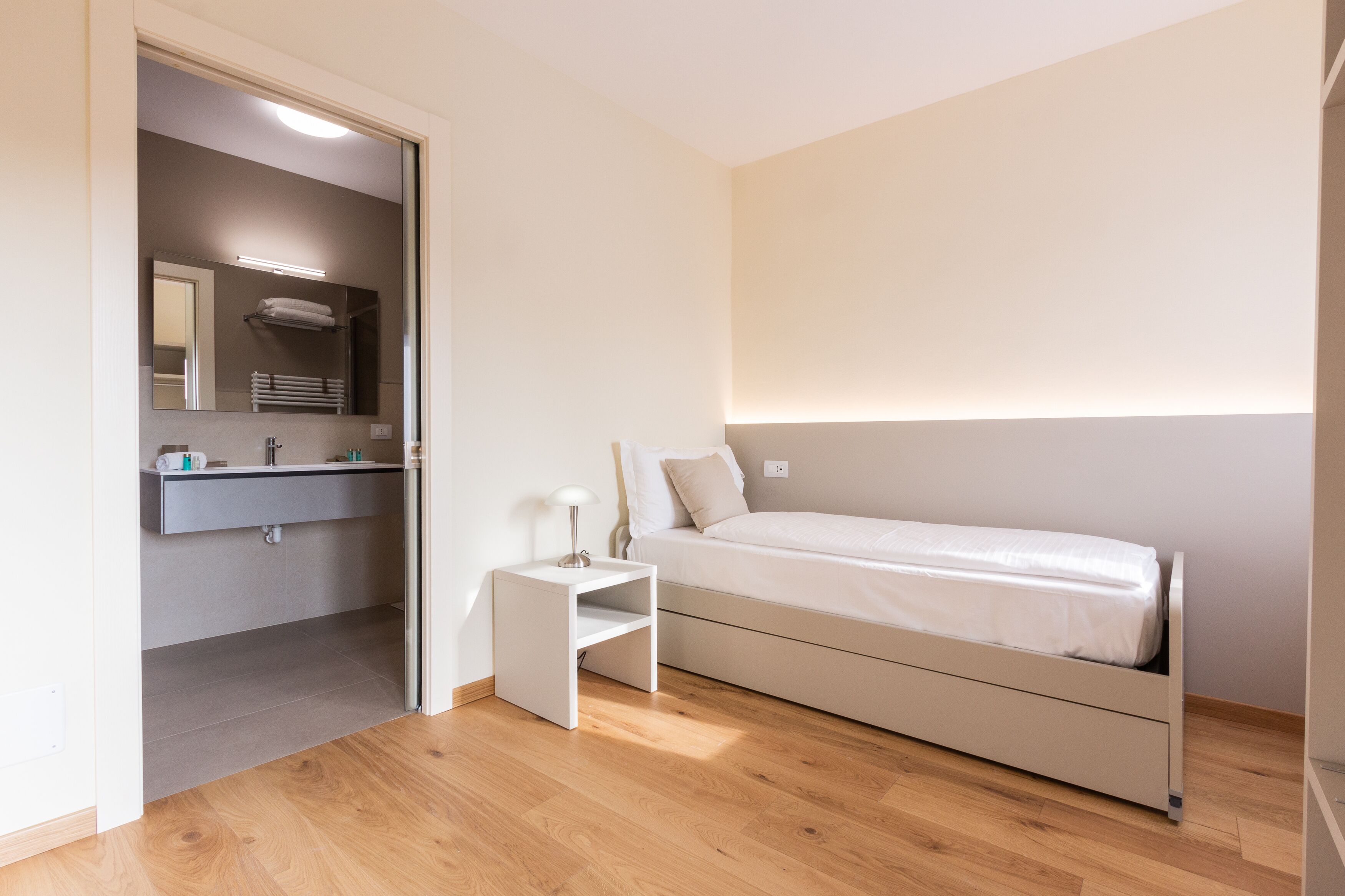 A bedroom with a bed, a nightstand, a mirror, and a bathroom visible through an open door.