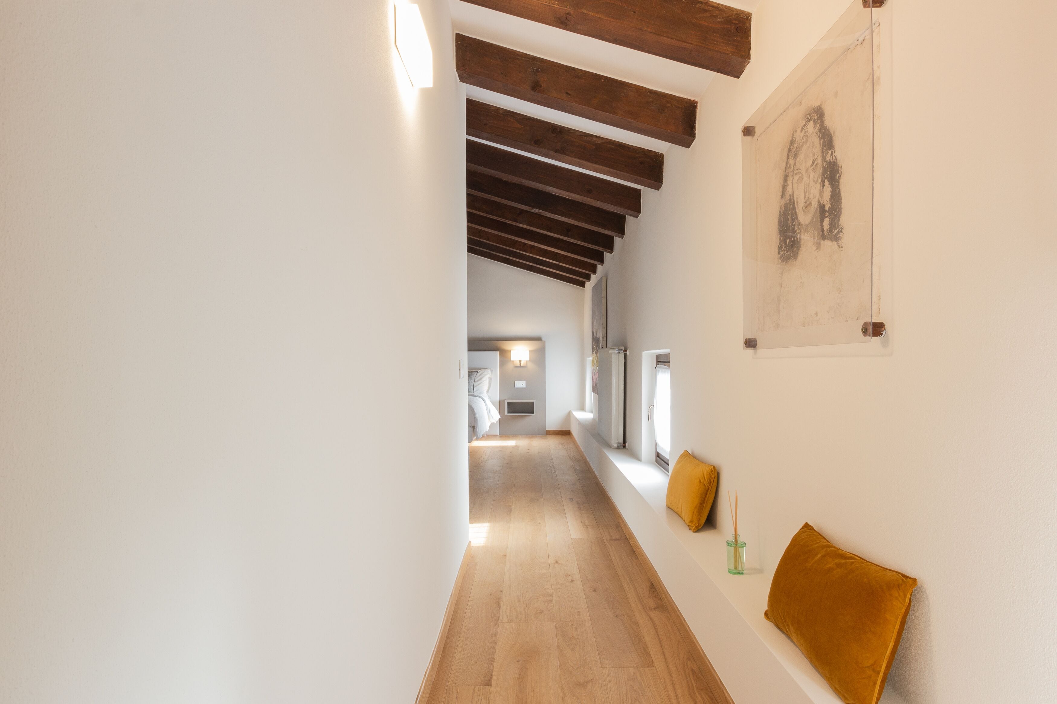 A hallway with wooden beams, a window with a yellow cushion, and a wall-mounted artwork.