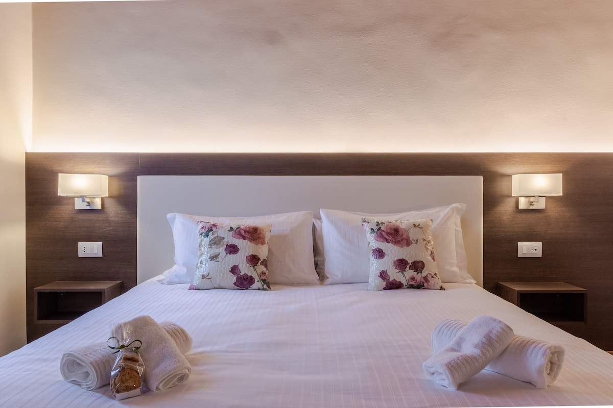 Un lit bien fait, agrémenté d’oreillers à motifs floraux, deux tables de chevet avec des lampes et des serviettes enroulées posées sur le lit.