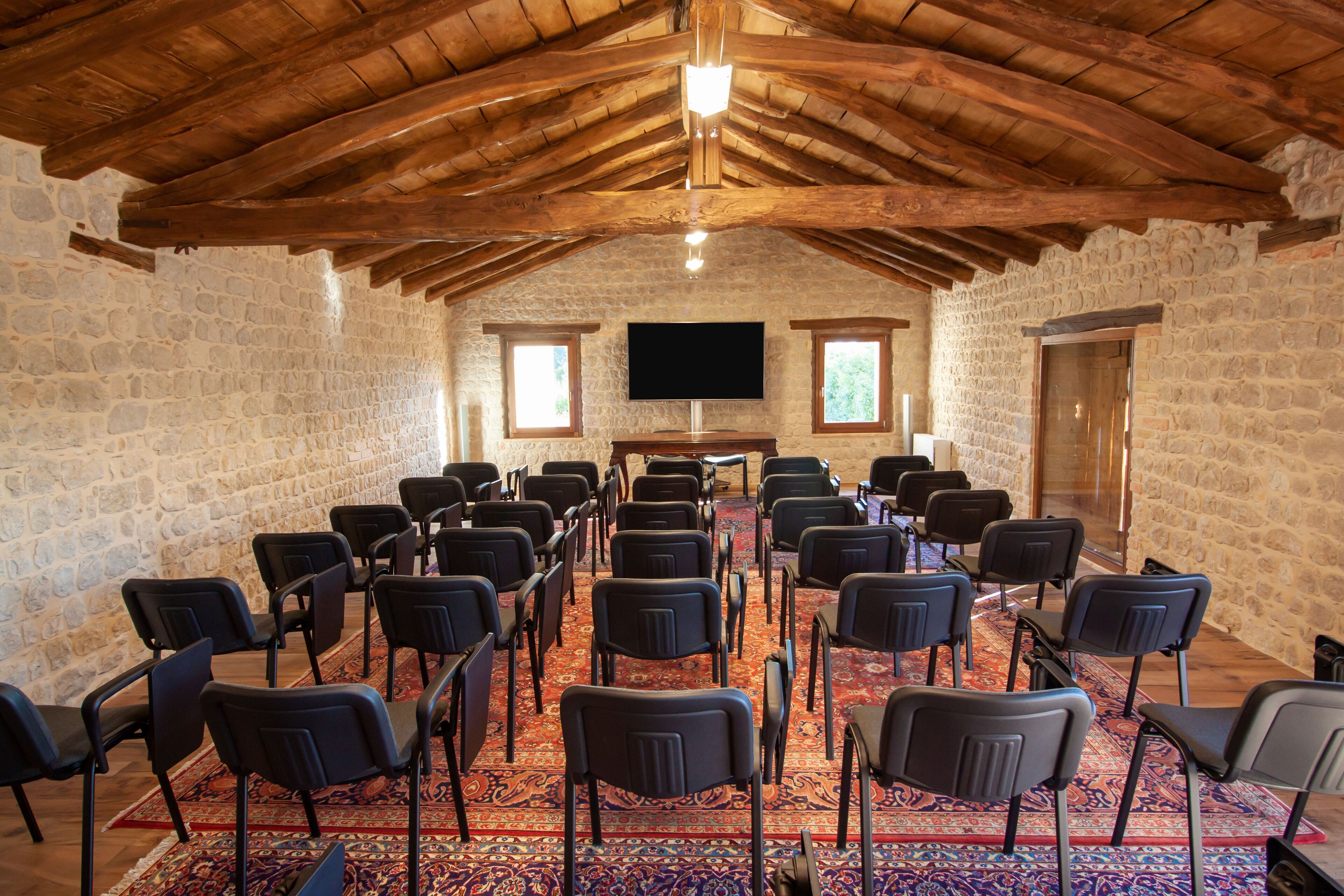 A conference room with stone walls, wooden ceiling beams, a large flat-screen TV, and rows of black chairs.