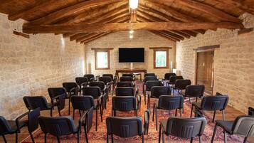 A conference room with stone walls, wooden ceiling beams, a large flat-screen TV, and rows of black chairs.