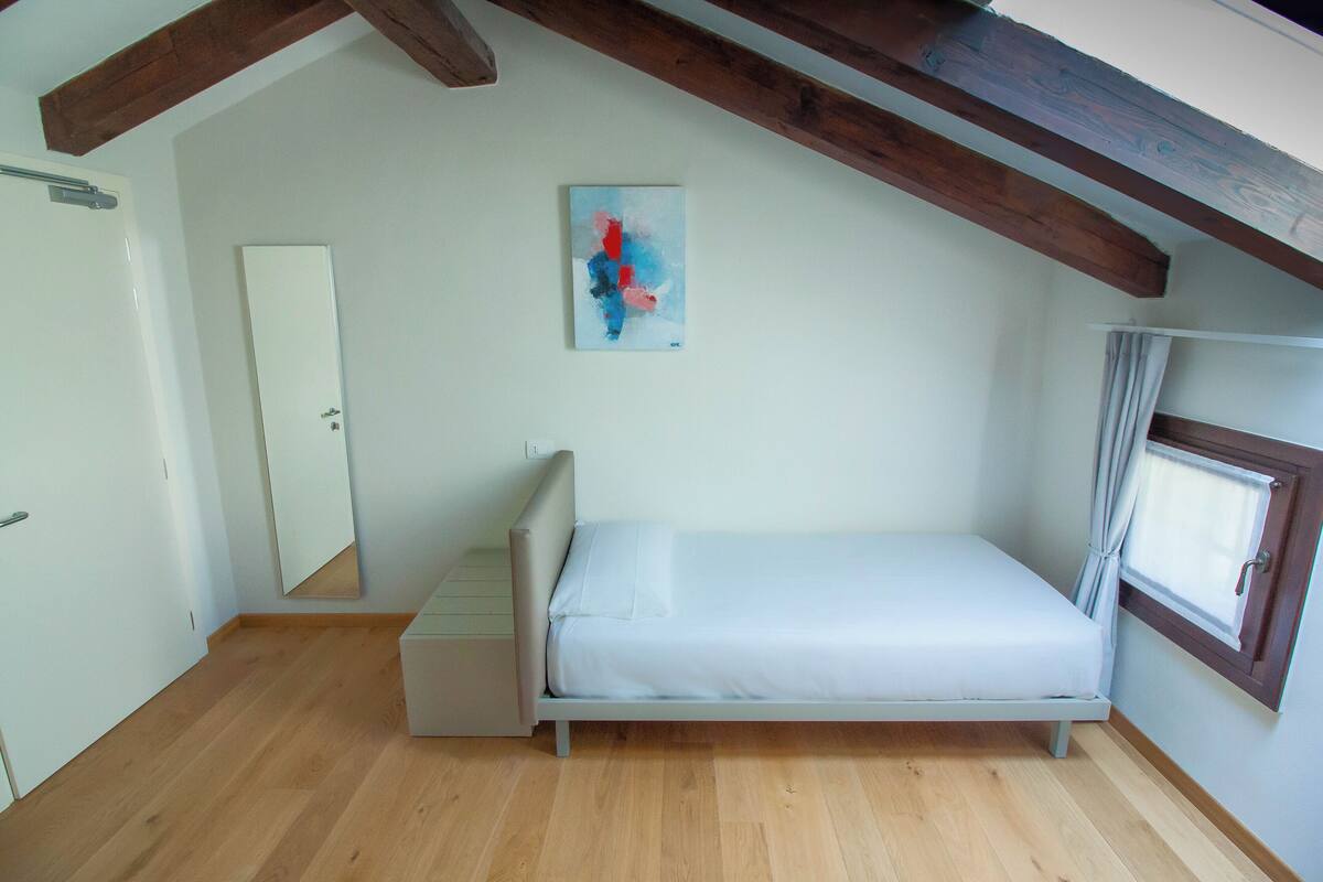 Une petite chambre avec un lit simple, un plafond en pente et des poutres en bois, une porte blanche, une fenêtre avec un rideau et un tableau accroché au mur.
