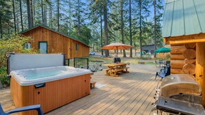 En træterrasse med jacuzzi, picnicbord og grillområde, omgivet af træer og hytter.