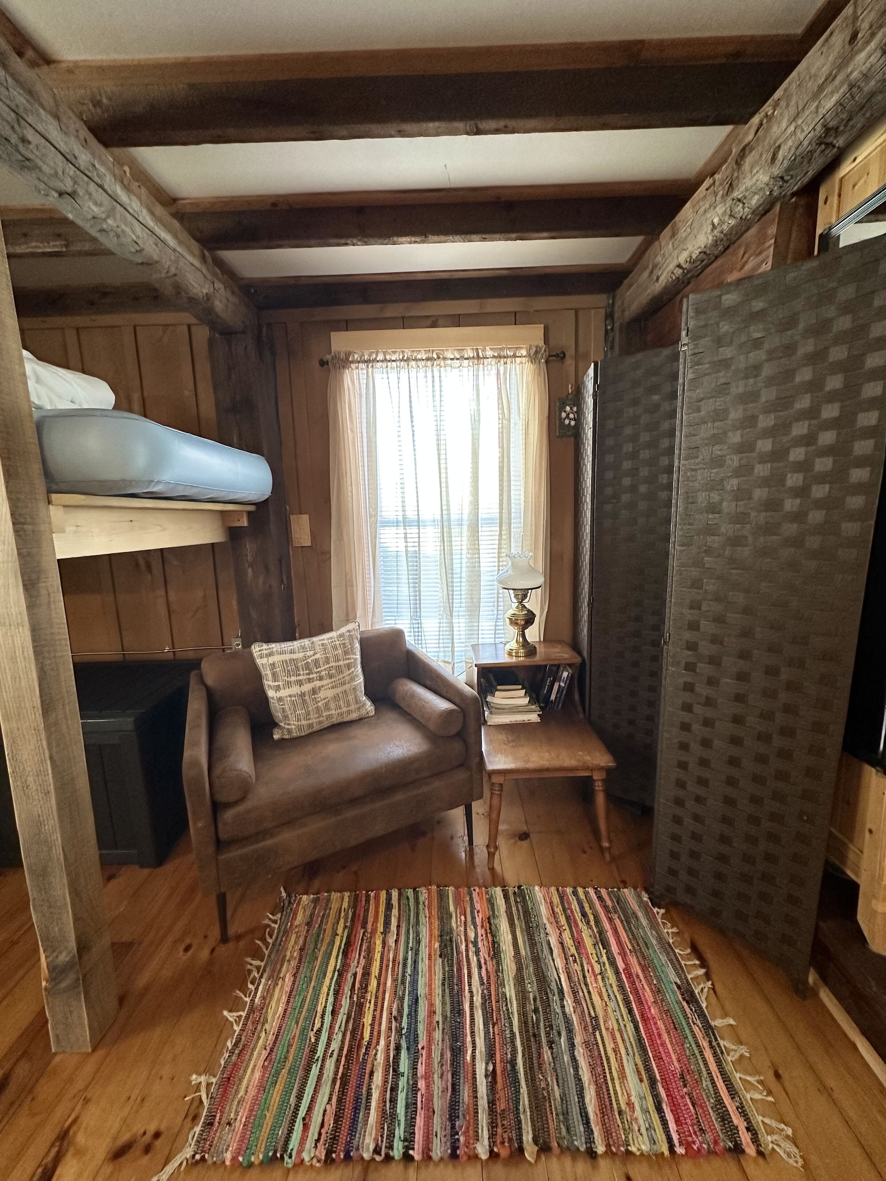 A cozy cabin interior with a wooden ceiling, a brown leather sofa, a colorful striped rug, and a bunk bed with a mattress on the left.
