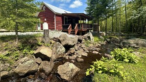 Un chalet rustique doté d’une terrasse en bois et d’un petit porche, entouré d’un ruisseau aux rives rocheuses et d’une végétation luxuriante.