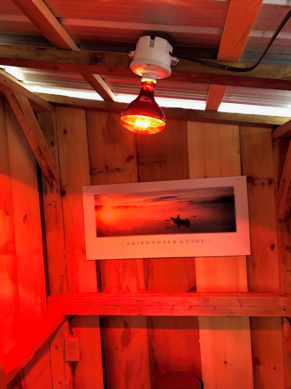 Interieur van een houten hutje met een plafondventilator, een lijst met een zonsondergangafbeelding en een zichtbare elektra-aansluiting.