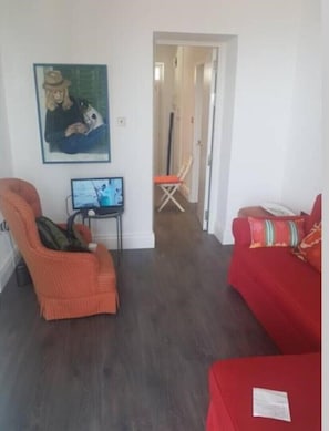 Una sala de estar con un sofá rojo, un sillón naranja, una mesita con un ordenador portátil y un cuadro en la pared.