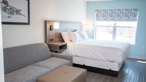 Un dormitorio moderno con un sofá gris, una cama con ropa blanca, una mesita de noche con una lámpara y una obra de arte con pájaros enmarcada en la pared.