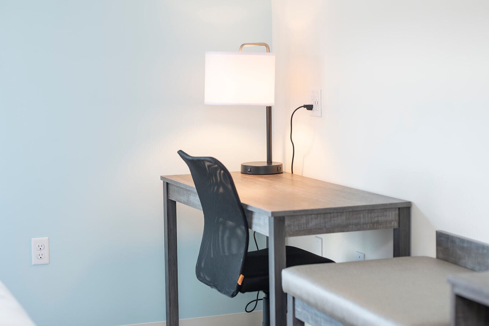 Ein kleiner, moderner Raum mit einem Holztisch, einem Stuhl, einer Tischlampe und einer Steckdose an der Wand.