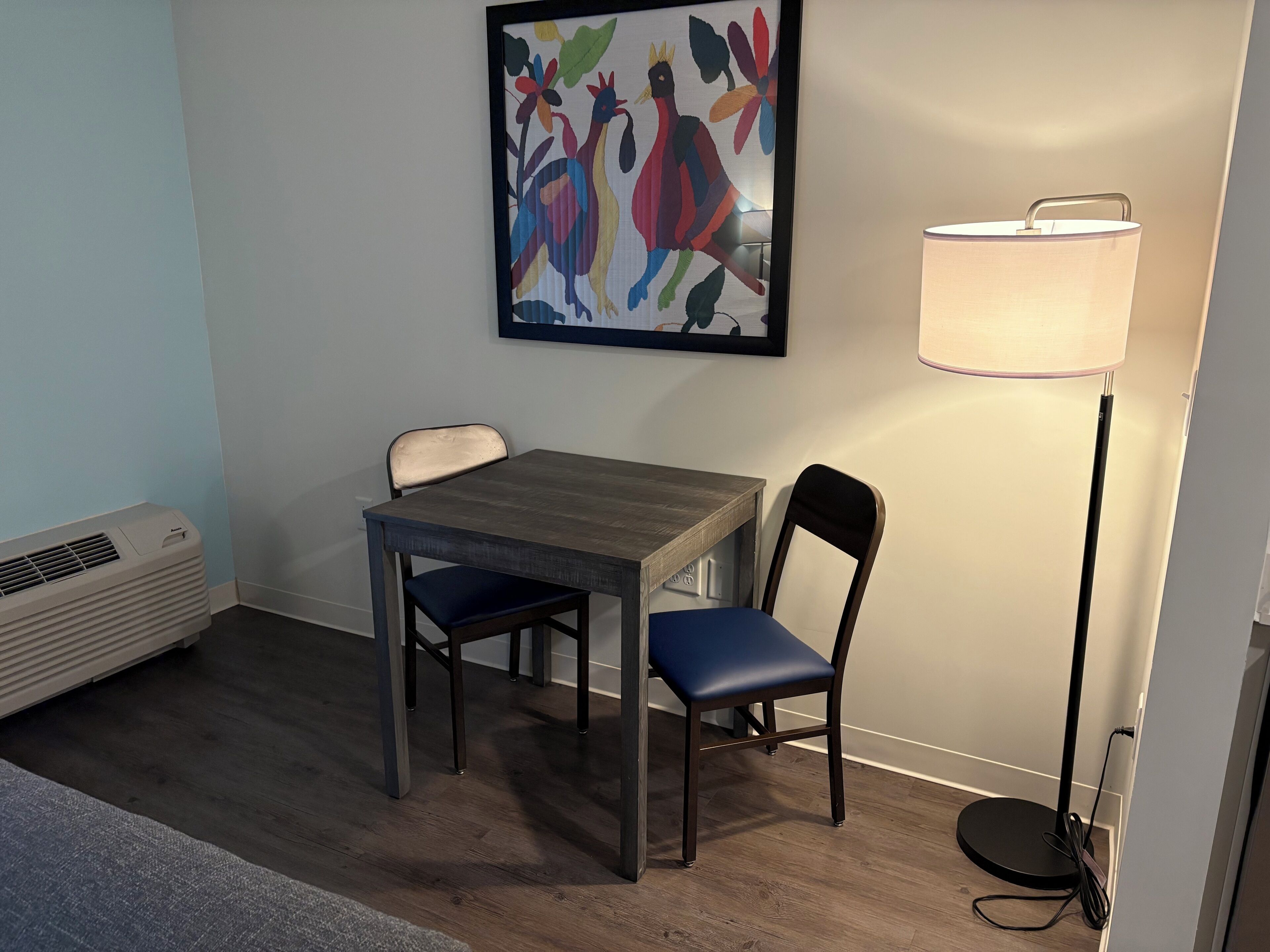 A room with a wooden table and two chairs, a floor lamp, and a colorful artwork on the wall.