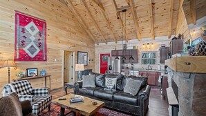 A cozy log cabin interior with a living room featuring a leather sofa, a checkered armchair, and a coffee table. The kitchen is visible with wooden cabinets and a stone fireplace. - Starway to Heaven Cabin at Red River Gorge  - Hot Tub - Great View (Frenchburg)