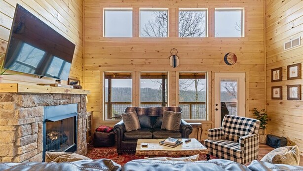 A cozy cabin interior with a stone fireplace, leather sofas, a checkered armchair, and a television mounted on the wall. - Starway to Heaven Cabin at Red River Gorge  - Hot Tub - Great View (Frenchburg)