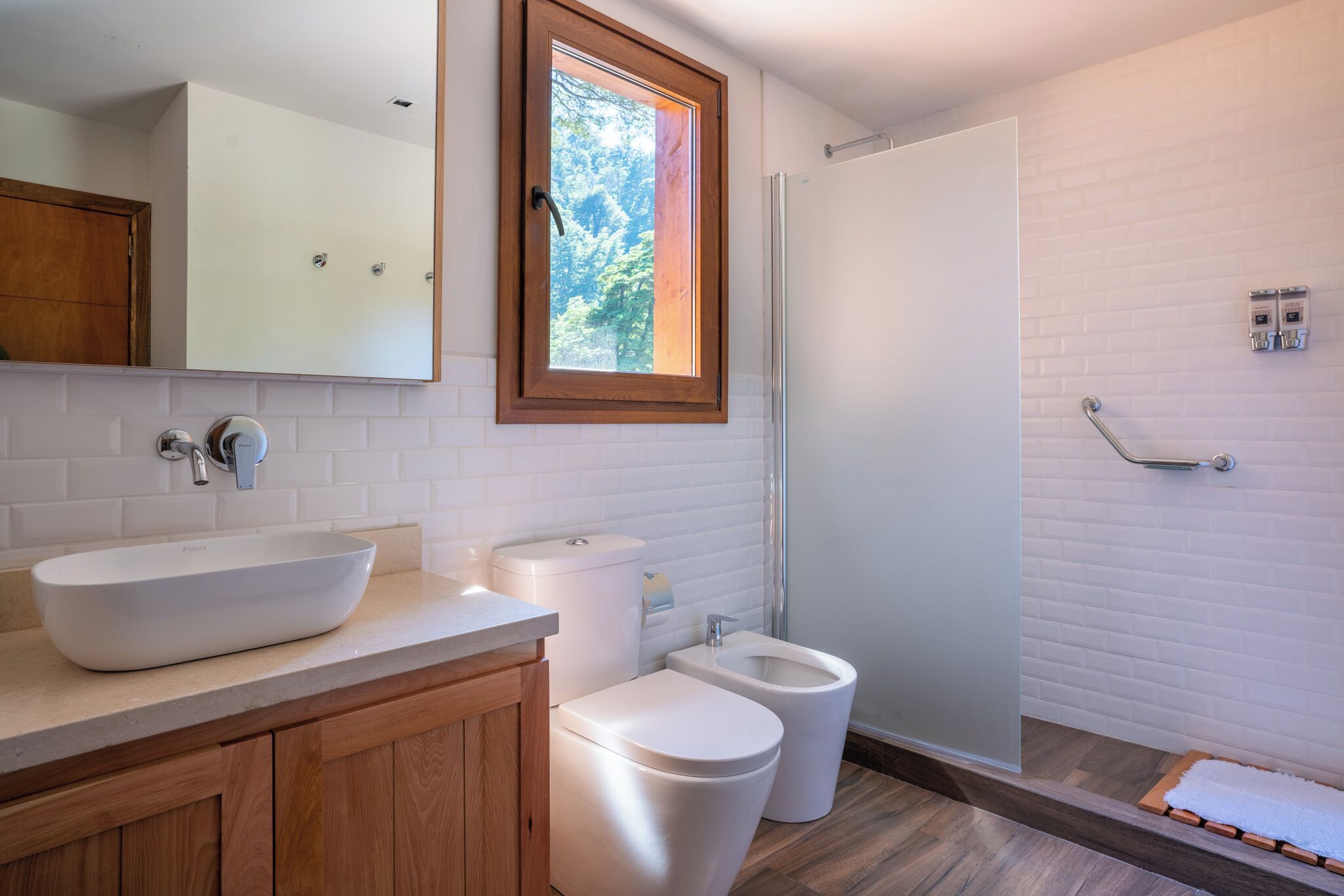A modern bathroom with a large white sink, a toilet, and a bidet. There is a wooden cabinet under the sink and a window above the toilet.