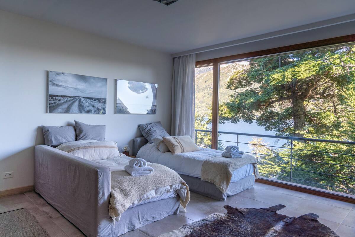 A bedroom with two beds, a large window with a view of trees and mountains, and artwork on the wall.