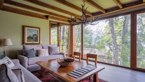 Amplia sala de estar con una gran mesa de madera, un sofá y vista a árboles a través de grandes ventanales.