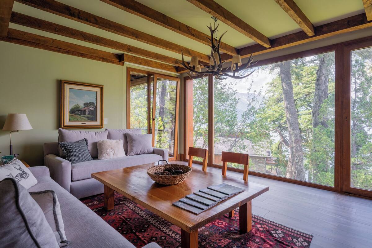 A spacious living room with a large wooden table, a sofa, and a view of trees through large windows.