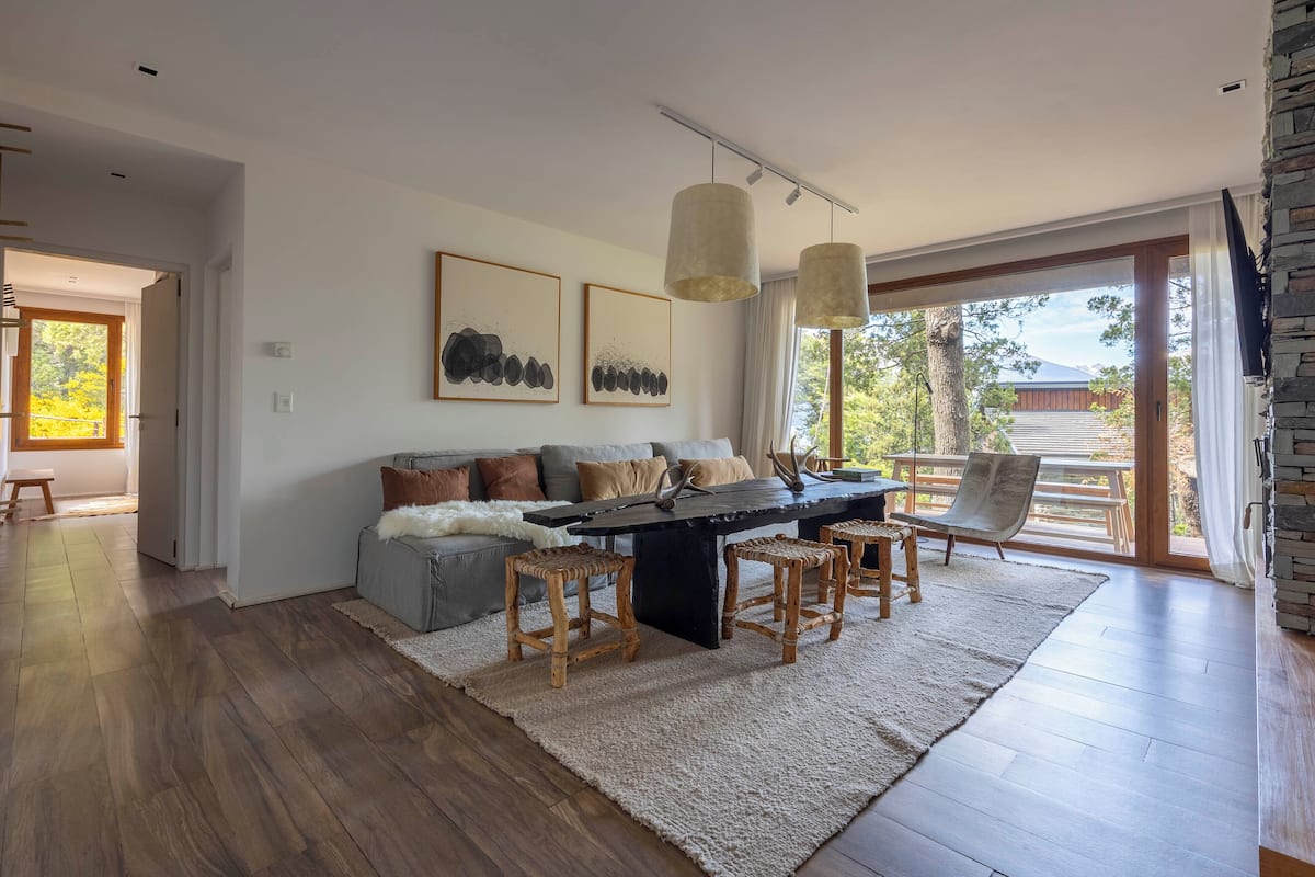 A modern living room with a sofa, coffee table, and wooden stools. There are two framed artworks on the wall, and a large window with a view of the outdoors.