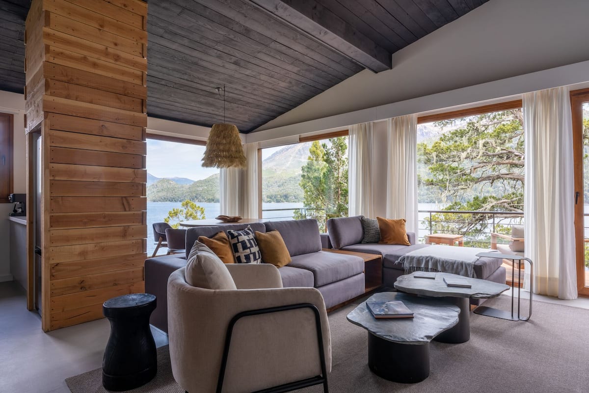 A modern living room with a large sofa, a coffee table, and a view of a lake and mountains.