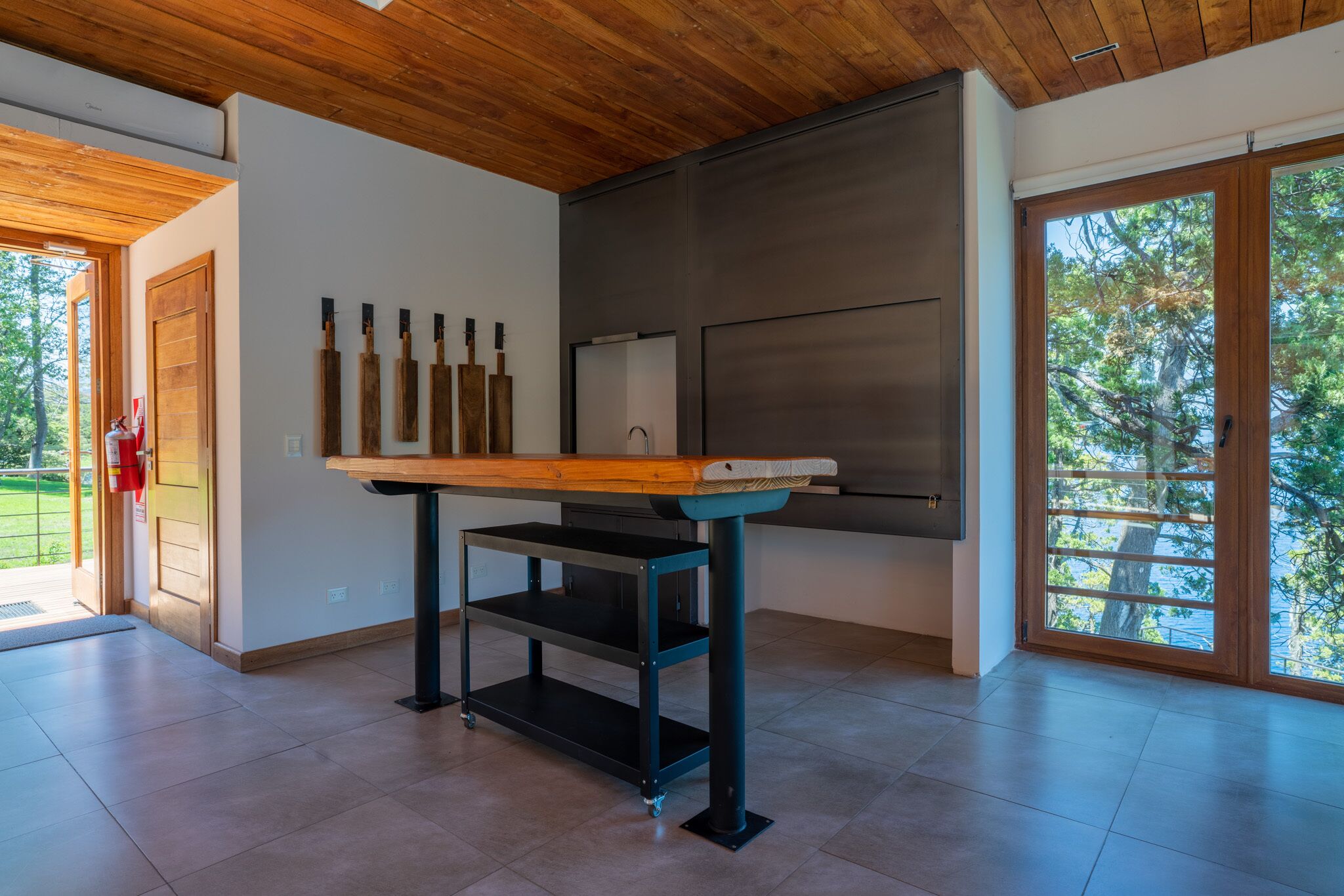 A modern kitchen with a wooden table, black metal shelves, and a large window with a view of trees.