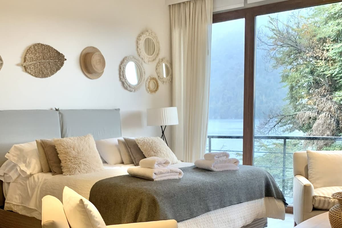 A modern bedroom with a large bed, a sitting area with a chair and a table, and a view of the outdoors through large windows.