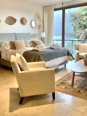 A modern bedroom with a large bed, a sitting area with a chair and a table, and a view of the outdoors through large windows. - Suites del Lago Bariloche  (San Carlos de Bariloche)