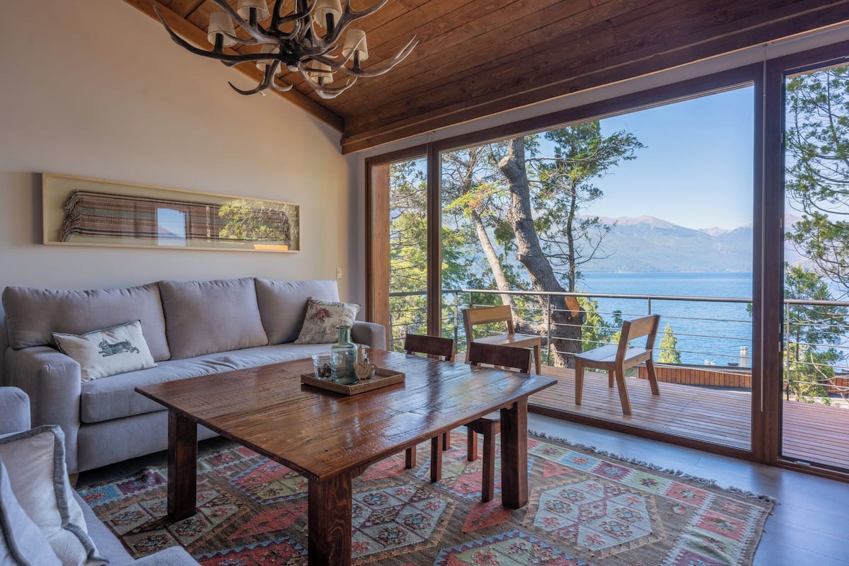 A cozy living room with a wooden coffee table, a sofa, and a view of a lake and mountains through large windows.