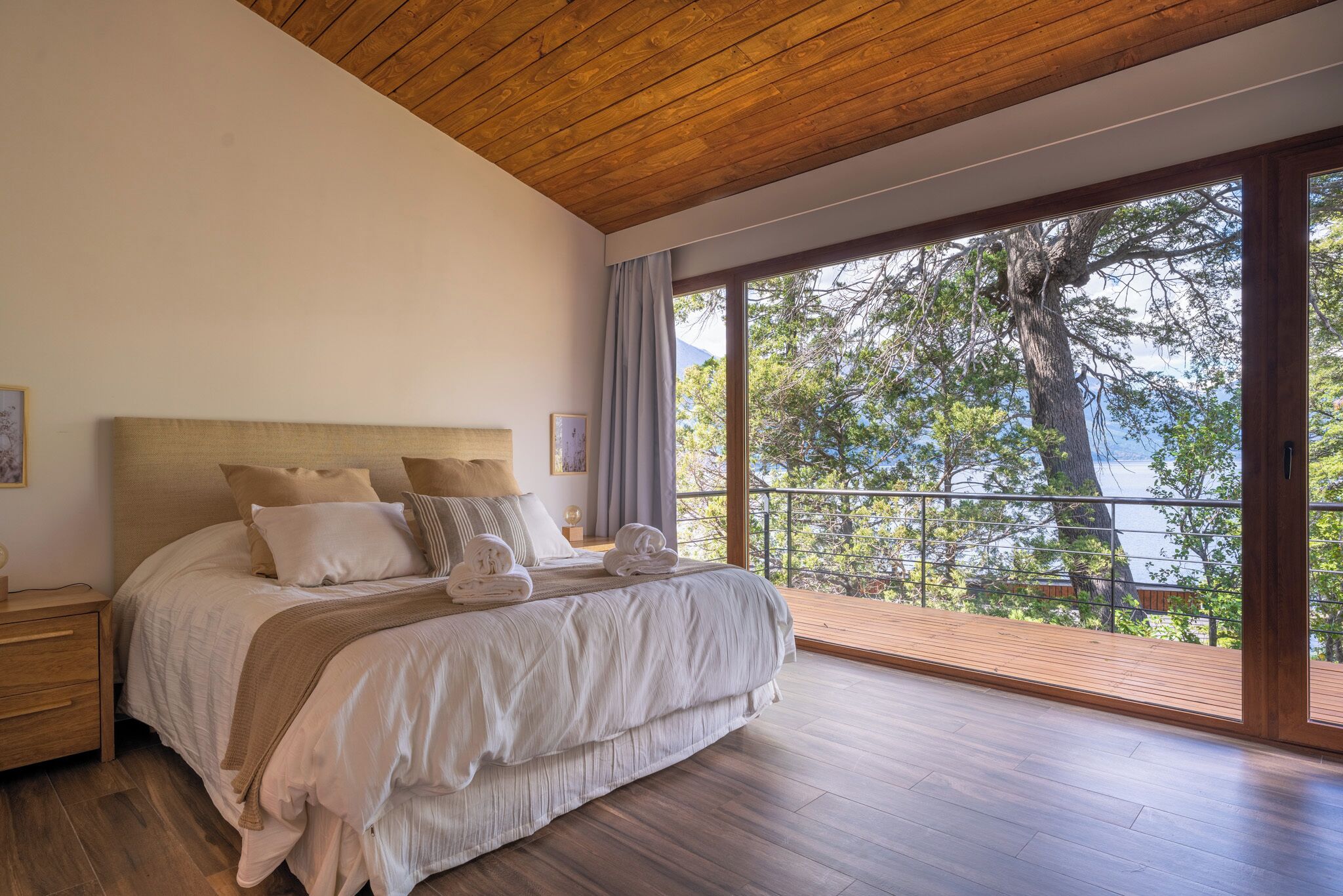A bedroom with a large bed, wooden flooring, and a balcony with a view of trees and a body of water.