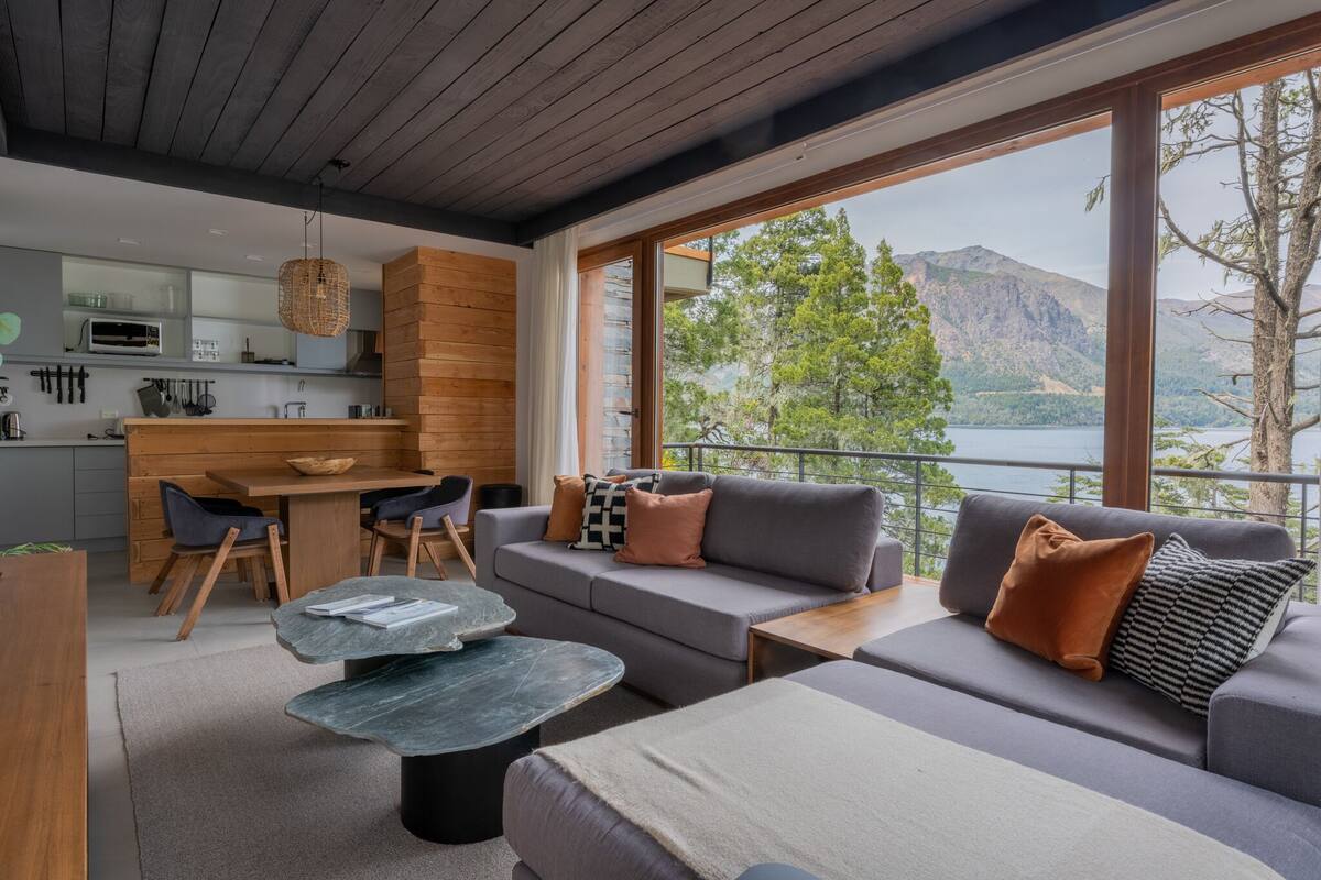 A modern interior with a kitchen, dining area, and a sofa set by a large window overlooking a lake and mountains.