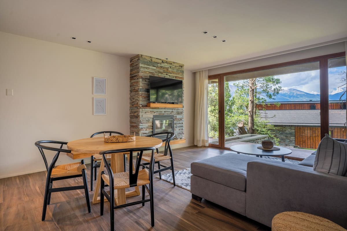 A modern living room with a stone fireplace, a dining area with a wooden table and chairs, and a large window offering a view of trees and mountains.