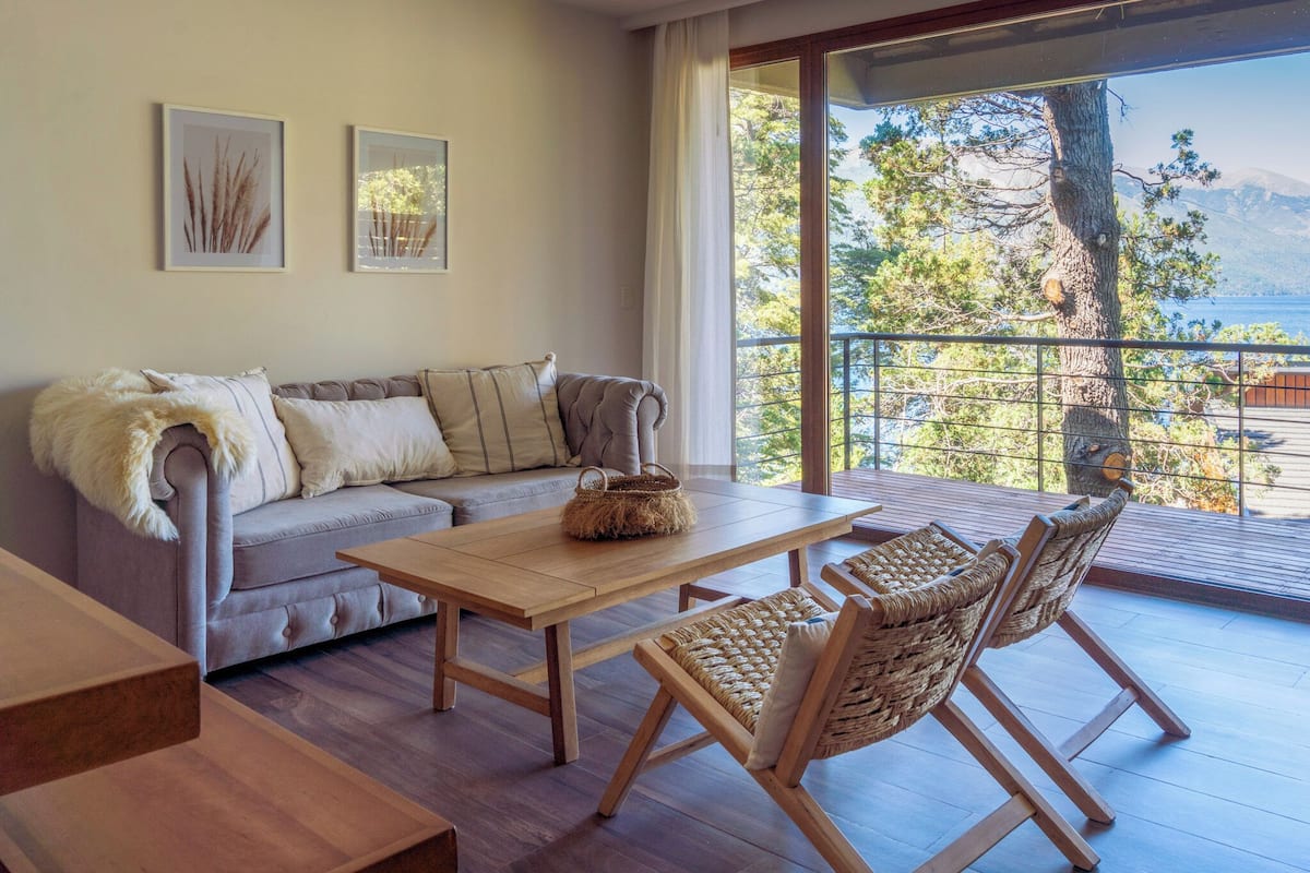 A cozy living room with a sofa, a wooden coffee table, and wicker chairs. There are framed pictures on the wall and a view of trees and mountains through a large window.