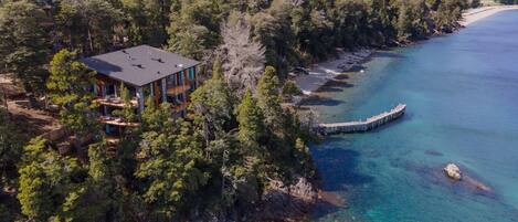 An aerial view of a coastal property with a large house surrounded by dense forest, a pier extending into the sea, and clear turquoise waters.