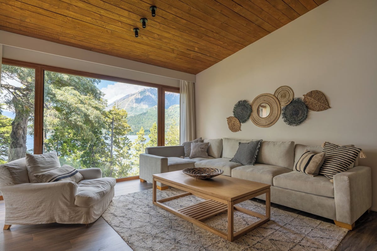 A modern living room with a wooden ceiling, large windows, a beige sofa, a wooden coffee table, and a view of mountains.