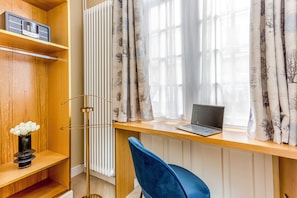 A room with a wooden desk, a laptop, a blue chair, a radiator, and a shelf with a vase of flowers.