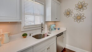 Una cocina con armarios blancos, un lavamanos de acero inoxidable, un lavavajillas y una ventana con persianas.