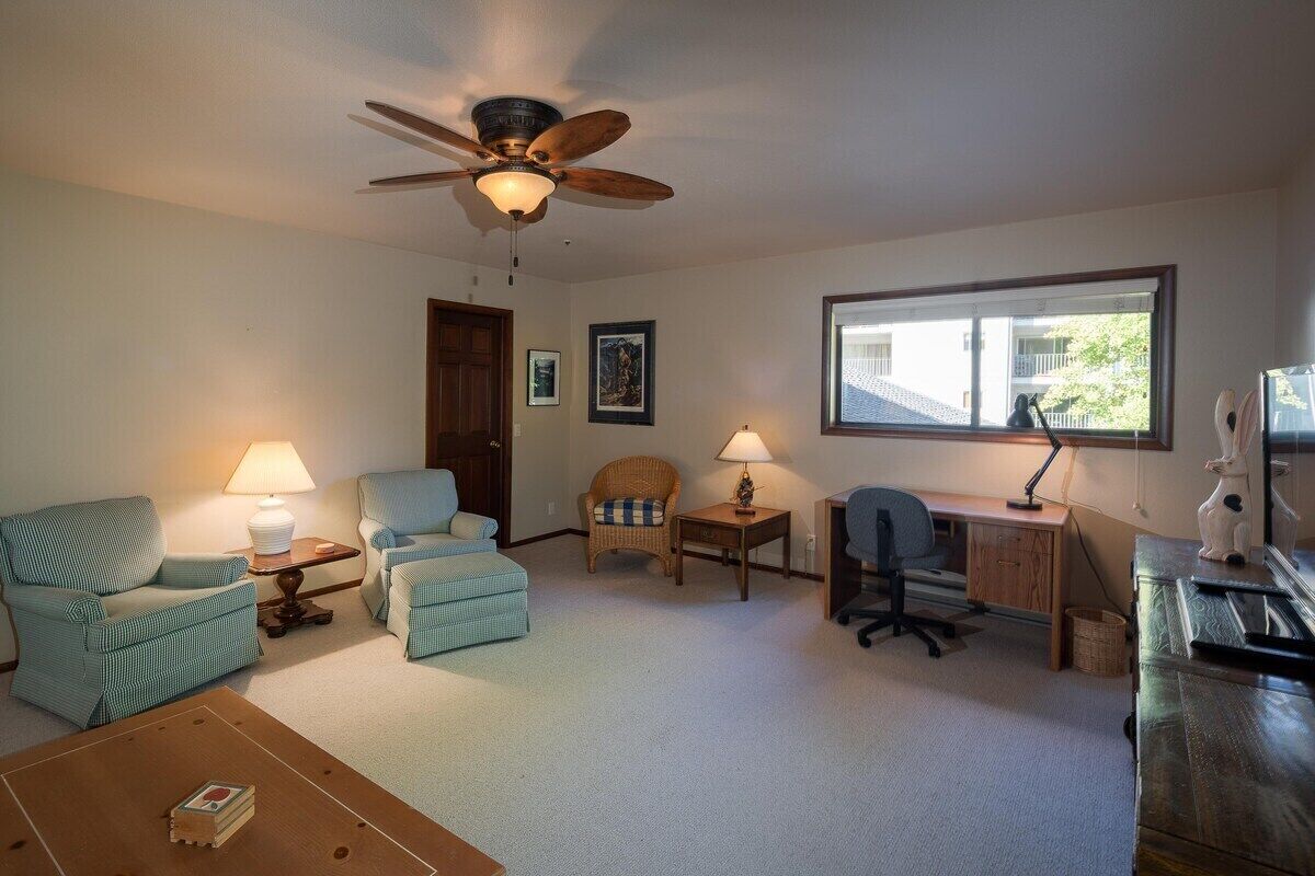 Un salon doté d’un ventilateur de plafond, de deux fauteuils, d’un bureau avec un ordinateur et d’une fenêtre laissant entrer la lumière naturelle.