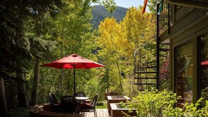 Un patio al aire libre con una mesa y sillas bajo un paraguas rojo, rodeado de abundante vegetación y una montaña al fondo.