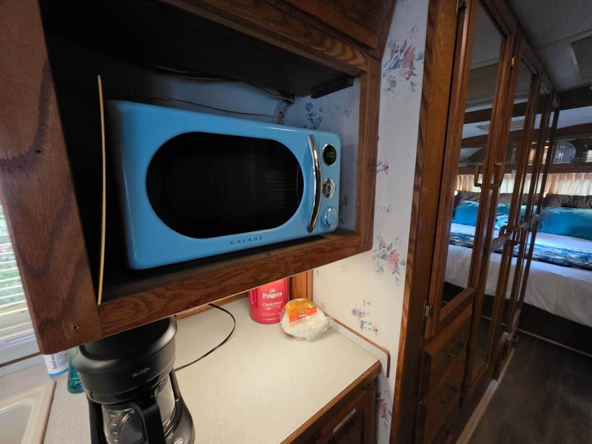 Angolo cottura con un forno a microonde blu su uno scaffale di legno, un contenitore rosso e un piano di lavoro bianco.